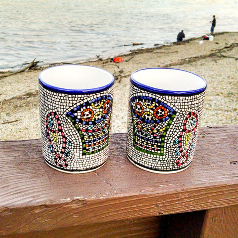 longdesc="2 Large Mosaic Glazed Ceramic Mugs that have two Red yellow Fishes and a blue and green basket containing round red and yellow pieces of bread loaves have been pattern pressed with a mosaic shape in black color. The almost square mosaic borders