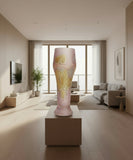 Vintage glassware standing on a small display behind a sunny livingroom with biege design and furniture. The long glass cup is Pastel Pink and has yellow metal blue swirls of hand painted stains. It has a Weizen shape for trapping the fuzzy bubbles in carbonated beverages. It is sort of like a microphone; that is wide at the top and thinner from 2/3rd of the highball glass to the bottom.