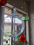 Large 7" Clear glass ornament with string lights against a window backdrop and twinkling fairy lights inside the orb, along with twinkling 3 inch green, and two red orbs.