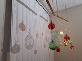 Colorful hanging christmas ornaments on a string against a shut white  window blinder with shadows of the ornaments .