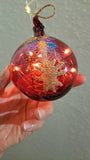 Decorative red blown glass ornament with christmas theme and fairy lights inside the onament; held by a hand against a neutral background