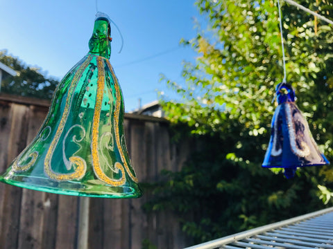 Decorative Glittered blown glass Green and Royal Blue bells hanging outdoors with a wooden fence and trees in the background.