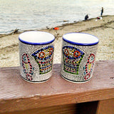 longdesc="2 Large Mosaic Glazed Ceramic Mugs that have two Red yellow Fishes and a blue and green basket containing round red and yellow pieces of bread loaves have been pattern pressed with a mosaic shape in black color. The almost square mosaic borders 